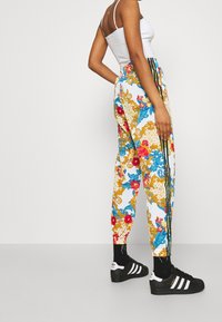 Färgglada mönstrade joggers i vitt med blå, röda och gyllene blommönster. Har svarta ränder längs sidorna. Bärs med svarta sneakers.