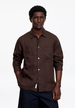 Homme à la peau foncée et aux cheveux courts et bouclés, portant une chemise marron à boutons et un pantalon sombre, debout avec une main dans la poche.