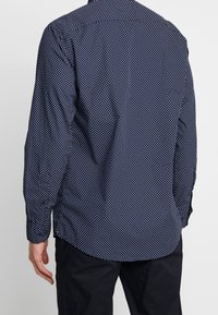 Camisa azul marino de botones con un estampado de lunares blancos, mangas largas y un dobladillo curvado. La tela parece suave y ligera.