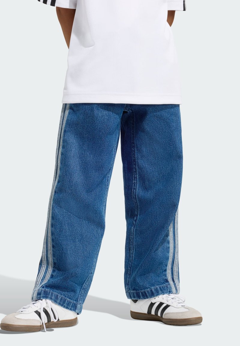 Jeans larghi in denim blu con strisce laterali bianche, caratterizzati da un taglio a gamba dritta, abbinati a sneakers bianche e nere.