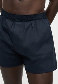 Άνδρας που φοράει σκούρες μπλε ελαστικές μποξεράκια, εμφανίζει τη μέση και το πάνω μέρος των μηρών του πάνω σε μονόχρωμο φόντο.