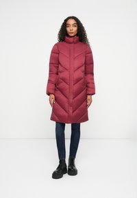 Giacca puffer bordeaux con colletto alto e motivo a chevron. Maniche lunghe e design a lunghezza media, abbinata a jeans blu scuro e stivali neri.