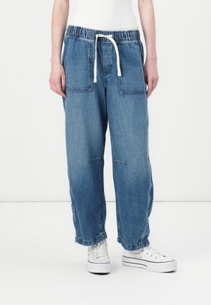 Person wearing loose blue denim pants with an elastic waistband and white drawstring, paired with white sneakers and a white top.