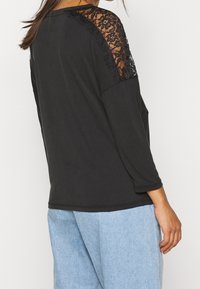 Maglietta nera a maniche lunghe con spalle decorate in pizzo, caratterizzata da una vestibilità morbida e una consistenza soffice, abbinata a pantaloni in denim azzurro chiaro.