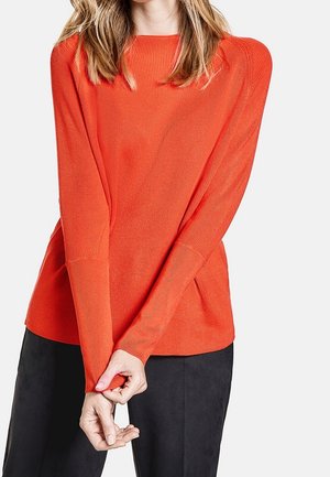 Femme portant un pull orange vif à manches longues avec une texture côtelée et un pantalon noir, mains croisées devant.