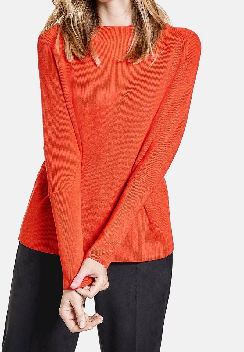 Femme portant un pull orange vif à manches longues avec une texture côtelée et un pantalon noir, mains croisées devant.