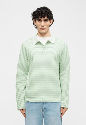 Νεαρό άτομο με κοντά σγουρά μαλλιά που φοράει μακρυμάνικο πόλο πουκάμισο με ριγέ μοτίβο σε ανοιχτό πράσινο και λευκό, και ανοιχτό μπλε τζιν, στέκεται μπροστά σε απλό φόντο.