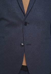Blazer blu navy con texture, dotato di due bottoni neri, rever a taglio e chiusura frontale, con una maglietta marrone chiaro visibile sotto.