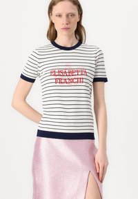 Top de manga corta a rayas en blanco y azul marino, que presenta una impresión de logotipo en rojo. Combinado con una falda metálica rosa con una abertura lateral.