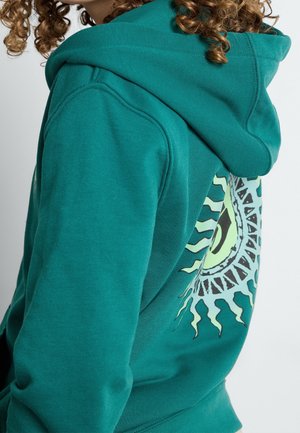 Lockighaarige Person trägt einen türkisfarbenen Hoodie mit einem großen stilisierten Auge und Sonnenstrahlen-Grafik auf dem Rücken.