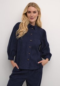 Mornarčeva plava košulja s dugim rukavima, s kragnom, napuhanim rukavima i zlatnim dugmetima. Glatka tkanina sa blagim teksturiranim završetkom.