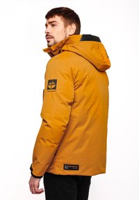 Uomo che indossa una giacca a vento gialla senape con loghi neri sulle maniche e sul retro, in piedi di lato contro uno sfondo bianco.