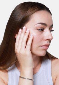 Modelo femenina con cabello largo castaño, vistiendo una blusa sin mangas de color gris claro, sostiene su rostro con una mano, mostrando la textura natural de la piel y una pulsera plateada.