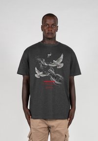 Grå bomulds t-shirt med et grafisk mønster af abstrakte kæder og duer, med rød tekst der står "FREEDOM" på brystet. Korte ærmer, afslappet pasform.