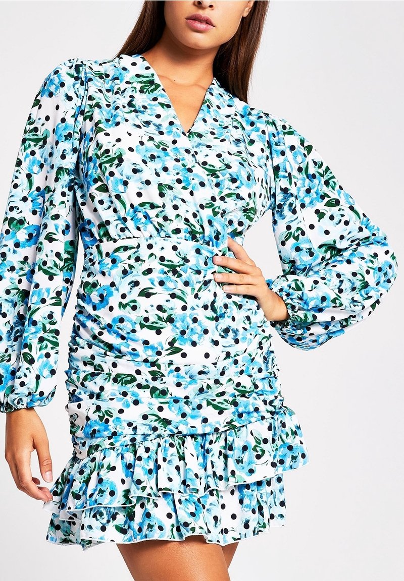 Žena ve světlých šatech s modrým květinovým a černým puntíkovaným vzorem, s dlouhými nafouklými rukávy, řaseným pasem a volánkovým lemem.