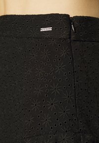 Gros plan sur un tissu noir avec des motifs floraux brodés et de petits trous, avec une fermeture éclair noire et une petite étiquette de marque blanche.