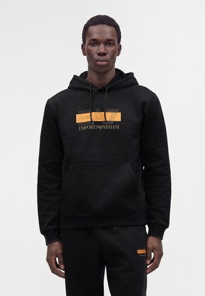 Jeune homme portant un sweat à capuche noir et un pantalon assorti avec le logo orange Emporio Armani EA7 sur la poitrine et la cuisse gauche.