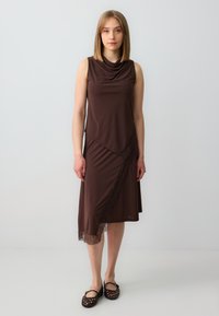Bruine mouwloze jurk met een gedrapeerde halslijn en een asymmetrische zoom, met kantaccenten aan de linkerkant. Model draagt zwarte gevlochten schoenen.
