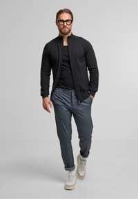 Schwarze Zip-Jacke, eng anliegendes schwarzes T-Shirt, strukturierte graue Hose und beigefarbene Sneakers mit weißen Akzenten. Das Model steht vor einem hellen Hintergrund.