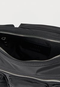 Fekete nejlon táska cipzáras nyílással, sima textúrával és ezüst cipzár részletekkel. A belsejében egy logófolt és anyag bélés látható.