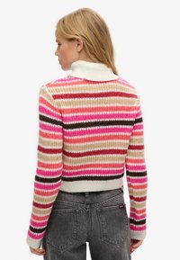 Pull rayé en rose, rouge, beige et marron. Texture en tricot avec un col haut et des poignets côtelés. Design ajusté, associé à un pantalon en jean gris.