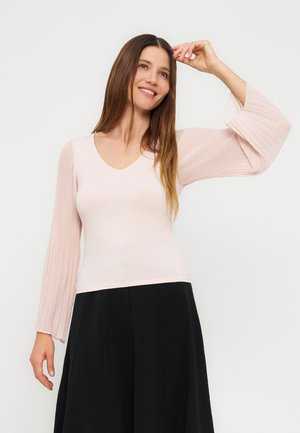 Kvinde med langt brunt hår iført en lyserød top med plisserede klokkeærmer og sorte vide bukser, smilende og med den ene hånd løftet.