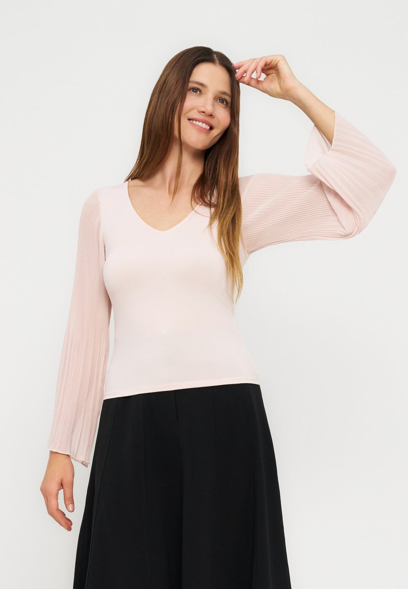 Γυναίκα με μακριά καστανά μαλλιά, που φοράει ανοιχτό ροζ μπλούζα με πλισέ μανίκια καμπάνες και μαύρο παντελόνι φαρδύ, χαμογελά και σηκώνει ένα χέρι.