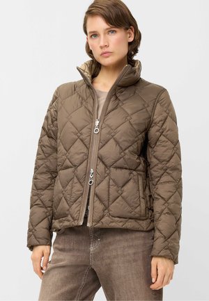 Woman wearing a brown quilted jacket with a high collar and double front zipper, paired with brown jeans against a plain light background.