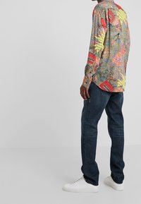 Långärmad skjorta i flerfärgad tyg med geometriska mönster och text, kombinerad med mörk denimjeans och vita sneakers.