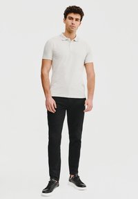 Męska koszulka polo w jasnym beżu, krótki rękaw, klasyczny kołnierzyk, zestawiona z czarnymi spodniami slim-fit i czarnymi butami sportowymi. Gładka tekstura.