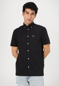 Camisa negra de manga corta con botones, con un bolsillo en el pecho y un pequeño logo de color, hecha de una tela texturizada. Lleva puesta con jeans desgastados de un azul claro.