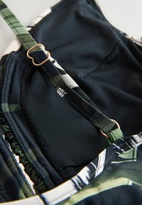 Juodos ir žalios spalvos maudymosi kostiumėlis su reguliuojamomis petnešėlėmis, papuoštas rožinio aukso sagtele ir tekstūruota medžiaga, suteikiančia papildomo elastingumo.