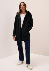 Veste en polaire noire avec capuche, fermeture éclair à l'avant et poches latérales. Associée à un jean bleu droit et des baskets blanches avec des accents noirs.