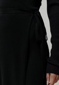 Schwarzes, geripptes Strickkleid mit einem gebundenen Taillendetail und langen Ärmeln. Der Stoff hat eine strukturierte Optik und eine schmale Passform.