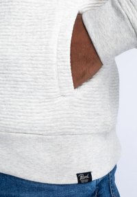 Ljusgrå texturerad sweatshirt med ribbat fåll, med en sidficka. Svart logotag sydd på undersidan. Denimjeans synliga nedanför.