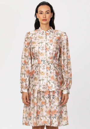 Femme portant une robe fleurie à manches longues ceinturée, avec des détails de boutons et un motif doux pêche, beige et vert sur fond clair.