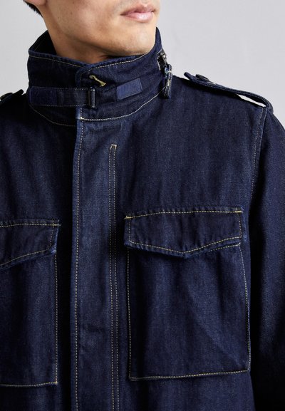 Denim jacket in dark blue, featuring a high collar, zipper, and large front pockets with yellow stitching and shoulder epaulettes.