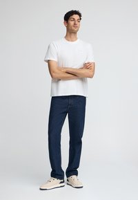 Witte katoenen t-shirt met een klein logo-patch, gecombineerd met donkerblauwe spijkerbroek. Sneakers zijn lichtbeige met marineblauwe accenten.