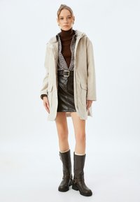 Abrigo beige con capucha sobre un suéter de cuello alto marrón y camisa de cuadros, con una falda corta de cuero negro. Completado con botas marrones hasta la rodilla.