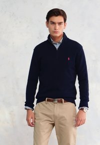 Pulover cu guler heringbone închis la culoare, dintr-un material tricotat moale, asortat cu o cămașă cu guler de culoare albastru deschis și pantaloni khaki. Detaliu discret cu logo pe piept.