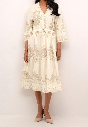 Kobieta w kremowej sukience midi w beżowe wzory kwiatowe, z marszczonym stanem ze sznurkiem, rękawami do łokcia oraz beżowych butach na szpic.