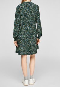 Blomklänning i mörkgrön med långa ärmar, med en rynkad rygg och en utsvängd nederdel. Buren med vita sneakers och grå strumpor.