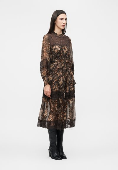 Γυναίκα με μακριά σκούρα μαλλιά, ντυμένη με καφέ λουλουδάτο δαντελένιο μίντι φόρεμα και μαύρες μπότες με τακούνι, στέκεται μπροστά σε απλό λευκό φόντο.