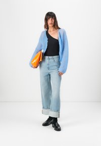Lichtblauwe, fluffy cardigan over een zwart top, gecombineerd met brede lichtdenim jeans met omgeslagen broekspijpen en zwarte loafers, terwijl een oranje clutch wordt vastgehouden.