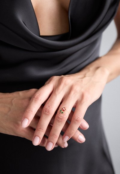 Anello d'oro con un piccolo design di rosa rossa e foglie verdi, su uno sfondo di tessuto scuro e liscio.