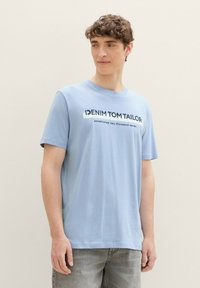 Šviesiai mėlynas medvilninis marškinėliai trumpomis rankovėmis, su juodu tekstu "DENIM TOM TAILOR" ir subtitru "įkurta du tūkstančiai septyni."