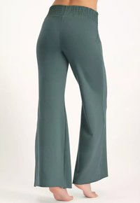 Nainen paljain jaloin, yllään korkea vyötärö, tilavat tummanvihreät housut, joissa on joustava vyötärönauha, näkyy takaa valkoista taustaa vasten.