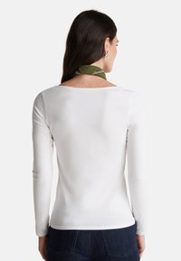 Camicia bianca a maniche lunghe con scollo a barca, texture liscia, design aderente, abbinata a una sciarpa verde con motivo annodata intorno al collo.