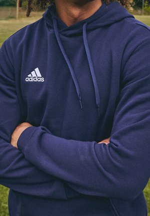 Osoba nosząca granatową bluzę Adidas z kapturem, z założonymi rękami, stojąca na zewnątrz na trawie, z rozmytymi drzewami w tle.