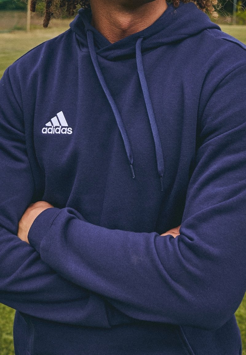 Personne portant un sweat à capuche Adidas bleu marine, les bras croisés, debout à l'extérieur sur l'herbe avec des arbres flous en arrière-plan.